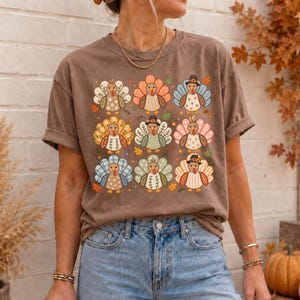 May include: A brown short-sleeve t-shirt featuring a grid of nine stylized turkeys. Each turkey has a unique patterned tail, including floral, plaid, and polka dot designs. Some turkeys wear small hats, and small autumn leaves are scattered around.