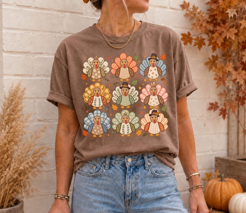 May include: A brown short-sleeve t-shirt featuring a grid of nine stylized turkeys. Each turkey has a unique patterned tail, including floral, plaid, and polka dot designs. Some turkeys wear small hats, and small autumn leaves are scattered around.