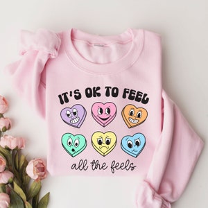 Pu&ograve; includere: Felpa rosa con il testo "It's OK to Feel all the Feels" e una fila di sei cuori di caramelle colorati con diverse espressioni facciali.