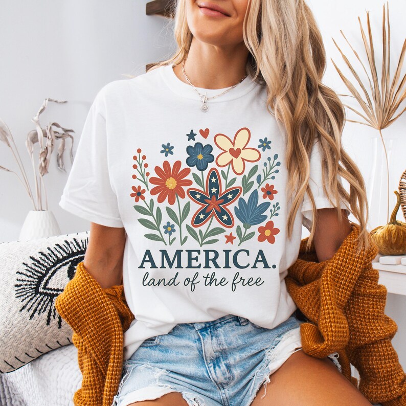 Peut inclure: T-shirt blanc avec un motif floral en rouge, blanc et bleu. Le motif comprend des &eacute;toiles et des fleurs. Le texte "AMERICA. land of the free" est imprim&eacute; sous le motif.