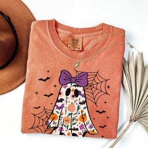 May include: A coral-coloured t-shirt with a ghost design, a purple bow, floral details, and a spiderweb. The shirt has bat and star graphics. A gold necklace and a brown hat are also visible.
