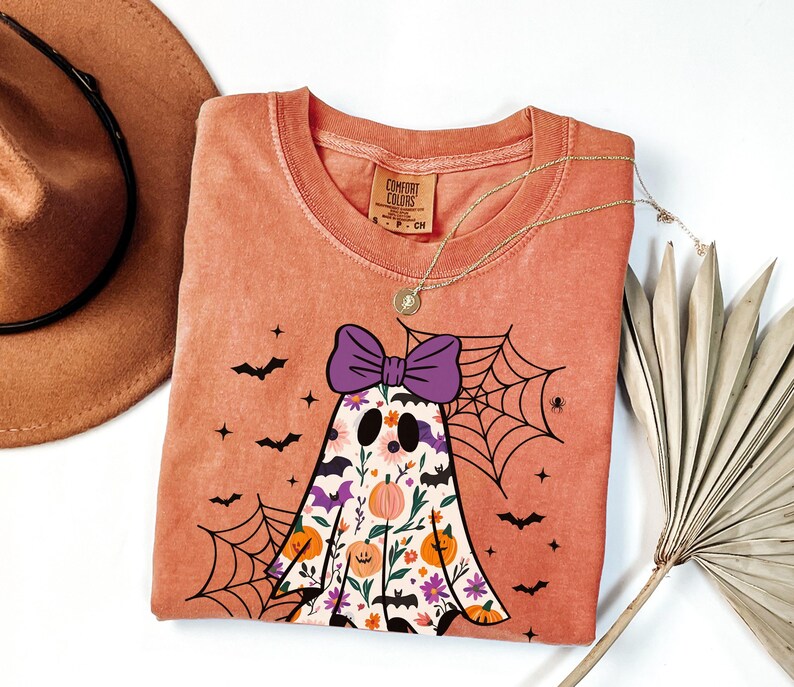 May include: A coral-coloured t-shirt with a ghost design, a purple bow, floral details, and a spiderweb. The shirt has bat and star graphics. A gold necklace and a brown hat are also visible.