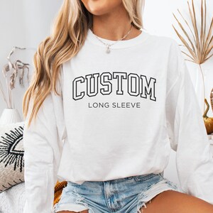 Op de afbeelding: Wit lange mouwen shirt met het woord "CUSTOM" in een zwarte, collegiale lettertype. Onder het woord "CUSTOM" staat de tekst "LONG SLEEVE" in een kleiner, zwart lettertype. Het shirt wordt gecombineerd met een spijkerbroek short.
