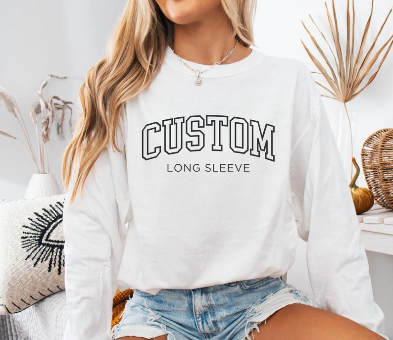Op de afbeelding: Wit lange mouwen shirt met het woord "CUSTOM" in een zwarte, collegiale lettertype. Onder het woord "CUSTOM" staat de tekst "LONG SLEEVE" in een kleiner, zwart lettertype. Het shirt wordt gecombineerd met een spijkerbroek short.