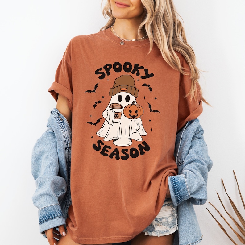 Op de afbeelding: Een roestkleurig T-shirt met een cartoon-spook dat een beanie draagt en een pompoen en een kopje koffie vasthoudt. De tekst "Spooky Season" is op het shirt gedrukt.
