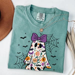 May include: Sea green t-shirt with a floral ghost design, purple bow, pumpkins, bats, and a spiderweb. A silver necklace with a pendant is draped across the shirt. The shirt is labelled "Comfort Colors".