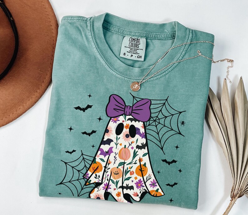 May include: Sea green t-shirt with a floral ghost design, purple bow, pumpkins, bats, and a spiderweb. A silver necklace with a pendant is draped across the shirt. The shirt is labelled "Comfort Colors".