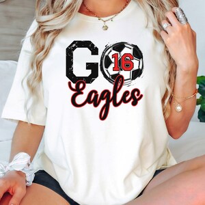May include: White t-shirt with a black and white football graphic and the text "GO Eagles" in red and black.
