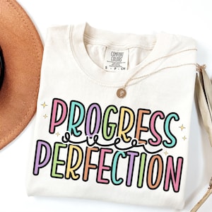 May include: Cream-colored t-shirt with the words "PROGRESS PERFECTION" in a colorful, outlined font. The shirt has a crew neck and short sleeves. A gold necklace and a brown hat are also in the image.