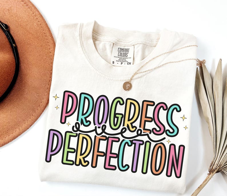 May include: Cream-colored t-shirt with the words "PROGRESS PERFECTION" in a colorful, outlined font. The shirt has a crew neck and short sleeves. A gold necklace and a brown hat are also in the image.