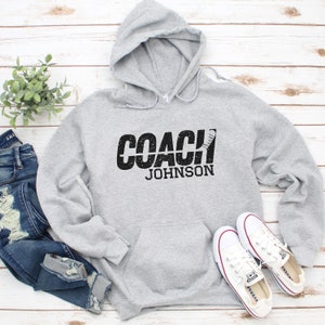 May include: Grey hooded sweatshirt with the text "COACH JOHNSON" in black with a hockey stick graphic.