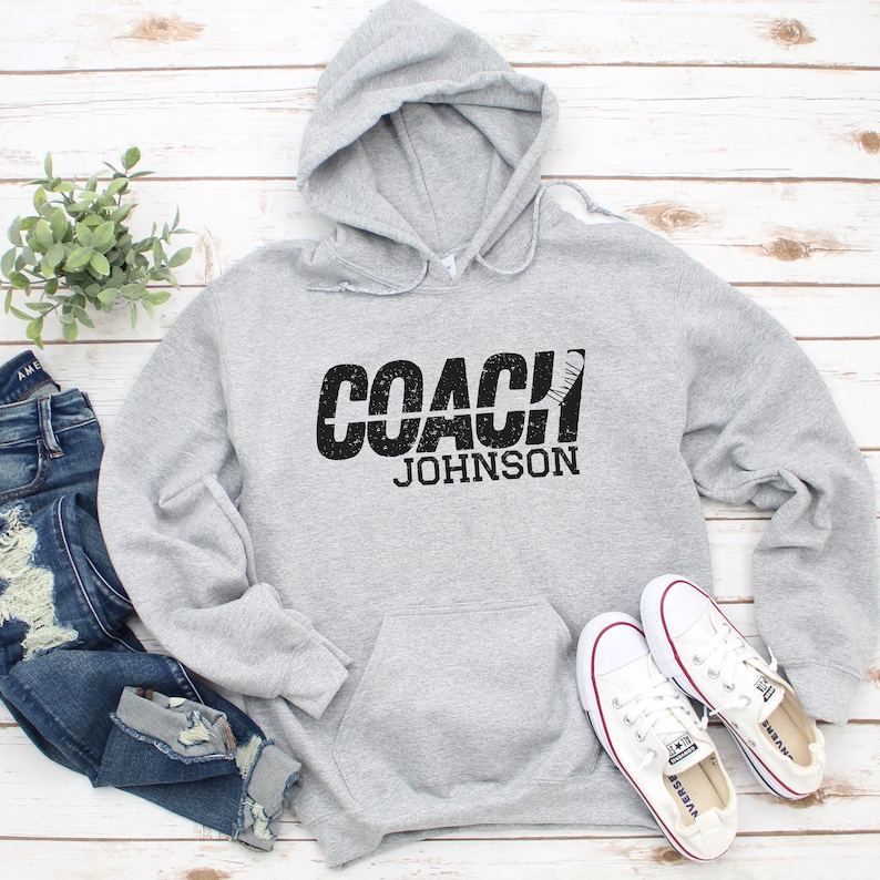 May include: Grey hooded sweatshirt with the text "COACH JOHNSON" in black with a hockey stick graphic.