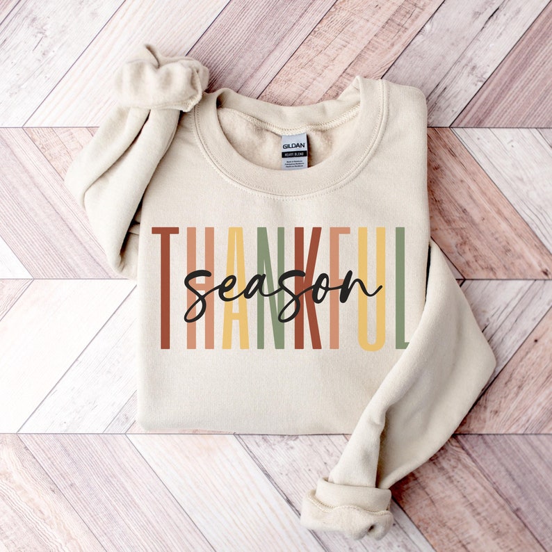 Puede incluir: Una sudadera beige con el texto "Thankful season" en rayas verticales de colores. El texto est&aacute; en un estilo de fuente moderno con un "season" escrito a mano en negro.