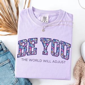 Pode incluir: Uma t-shirt roxo-claro com o texto BE YOU em letras grandes de bloco preenchidas com um padr&atilde;o floral colorido, seguido pelo texto THE WORLD WILL ADJUST em letras mais pequenas de cor roxo-escuro. A t-shirt tem uma etiqueta Comfort Colors.
