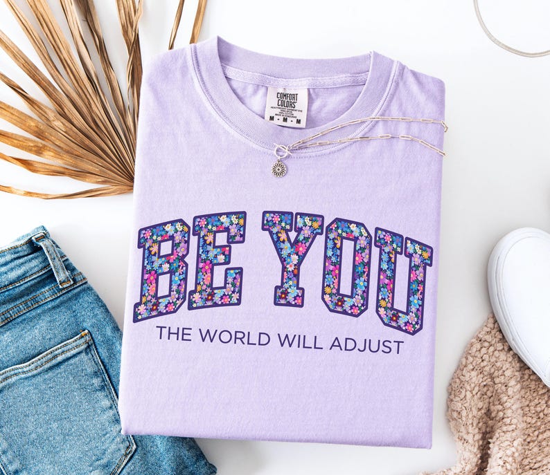 Pode incluir: Uma t-shirt roxo-claro com o texto BE YOU em letras grandes de bloco preenchidas com um padr&atilde;o floral colorido, seguido pelo texto THE WORLD WILL ADJUST em letras mais pequenas de cor roxo-escuro. A t-shirt tem uma etiqueta Comfort Colors.