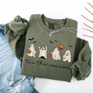 May include: An olive green sweatshirt with a crew neck featuring a Halloween-themed design. The graphic includes four cartoon ghosts, bats, and pumpkins. The phrase "tis the season" is printed below the ghosts. A silver necklace is draped on the sweatshirt.