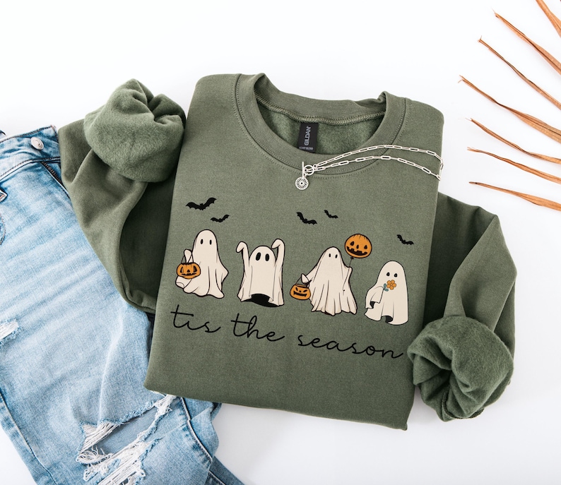 May include: An olive green sweatshirt with a crew neck featuring a Halloween-themed design. The graphic includes four cartoon ghosts, bats, and pumpkins. The phrase "tis the season" is printed below the ghosts. A silver necklace is draped on the sweatshirt.