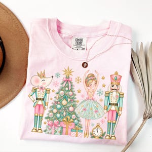May include: A light pink t-shirt featuring a graphic print of a mouse in a crown, a decorated Christmas tree, a ballerina, and a nutcracker. The design includes pastel-colored ornaments, gifts, snowflakes, and a clock. The shirt has a Comfort Colors label.