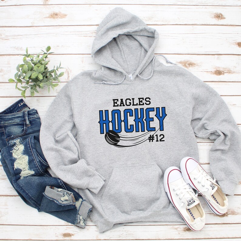 K&ouml;nnte beinhalten: Grauer Kapuzenpullover mit dem Text "Eagles Hockey #12" in blauer und schwarzer Schrift. Ein Hockeypuck und ein Schl&auml;ger sind im Design abgebildet.