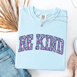 May include: A light blue Comfort Colors t-shirt with the words "BE KIND" in a floral design. A silver necklace with a small pendant rests on the shirt. The shirt is paired with blue jeans and a white sneaker.