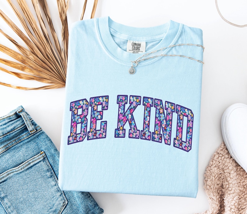 May include: A light blue Comfort Colors t-shirt with the words "BE KIND" in a floral design. A silver necklace with a small pendant rests on the shirt. The shirt is paired with blue jeans and a white sneaker.