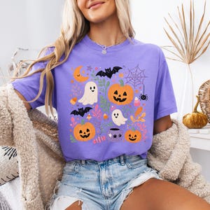 Może przedstawiać: Fioletowy t-shirt z kr&oacute;tkim rękawem z kolorowym nadrukiem graficznym o tematyce Halloween. Wz&oacute;r zawiera pomarańczowe dynie, białe duszki, czarne nietoperze, pajęczynę z pająkiem, p&oacute;łksiężyc oraz r&oacute;żnorodne liście w jesiennych kolorach.