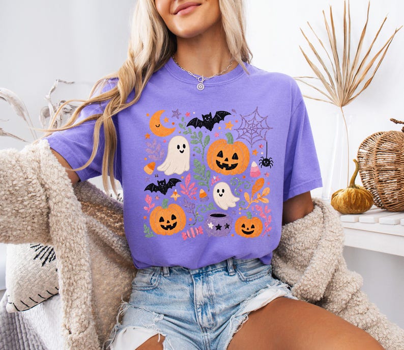Może przedstawiać: Fioletowy t-shirt z kr&oacute;tkim rękawem z kolorowym nadrukiem graficznym o tematyce Halloween. Wz&oacute;r zawiera pomarańczowe dynie, białe duszki, czarne nietoperze, pajęczynę z pająkiem, p&oacute;łksiężyc oraz r&oacute;żnorodne liście w jesiennych kolorach.