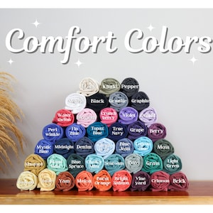Op de afbeelding: Een display van opgerolde t-shirts in verschillende kleuren, waaronder wit, zwart en blauw. De afbeelding toont de tekst "Comfort Colors" in een decoratief lettertype, waarbij elk shirt is gelabeld met zijn kleurnaam. De shirts zijn in een piramidevorm gestapeld.