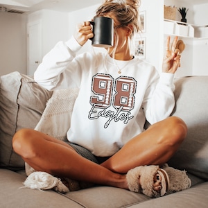 May include: White sweatshirt with a brown football number 98 and the word "Eagles" in black script.