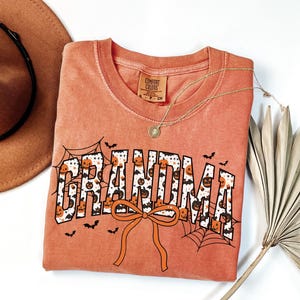 May include: Orange Comfort Colors t-shirt with the word "GRANDMA" in a Halloween-themed design. The letters are filled with pumpkins and bats, with spiderwebs and a bow. A gold necklace and a brown hat are also in the image.