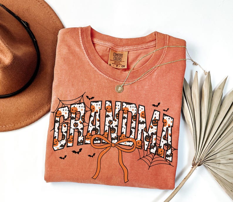 May include: Orange Comfort Colors t-shirt with the word "GRANDMA" in a Halloween-themed design. The letters are filled with pumpkins and bats, with spiderwebs and a bow. A gold necklace and a brown hat are also in the image.