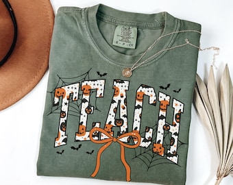Può includere: T-shirt verde oliva con la scritta "TEACH" in un design a tema Halloween. Le lettere sono riempite con motivi di zucche e stelle, circondate da ragnatele, pipistrelli e un fiocco arancione. Una collana d'argento con un ciondolo è drappeggiata sulla maglietta.