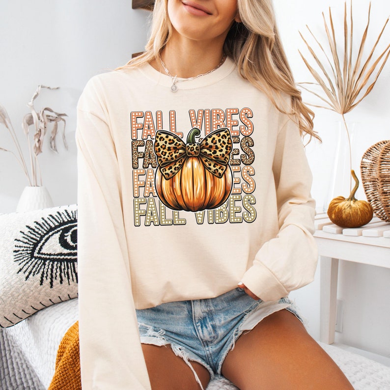 May include: Cream-coloured long-sleeve shirt with a pumpkin graphic and the text "FALL VIBES". The pumpkin has a leopard print bow. The shirt is a fall-themed apparel item.