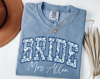 May include: A blue Comfort Colors t-shirt with the word "BRIDE" in a floral design, and the name "Mrs. Allen" underneath. A gold necklace with a pendant is draped across the shirt. A brown hat and dried leaves are in the background.