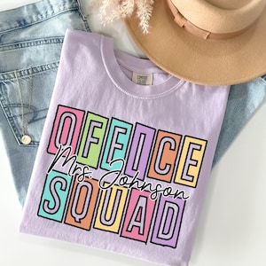 May include: A lilac-colored t-shirt with the words "OFFICE SQUAD Mrs. Johnson" in a colorful, block-letter design. The shirt is folded on top of blue jeans, with a tan hat and a gold necklace visible.