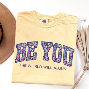 Pode incluir: Uma t-shirt amarelo-p&aacute;lido com o texto BE YOU em letras grandes e em bloco preenchidas com um padr&atilde;o floral colorido, seguido pelo texto THE WORLD WILL ADJUST numa fonte sans-serif azul-escura, com uma etiqueta da marca Comfort Colors.