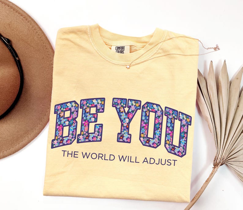 Pode incluir: Uma t-shirt amarelo-p&aacute;lido com o texto BE YOU em letras grandes e em bloco preenchidas com um padr&atilde;o floral colorido, seguido pelo texto THE WORLD WILL ADJUST numa fonte sans-serif azul-escura, com uma etiqueta da marca Comfort Colors.