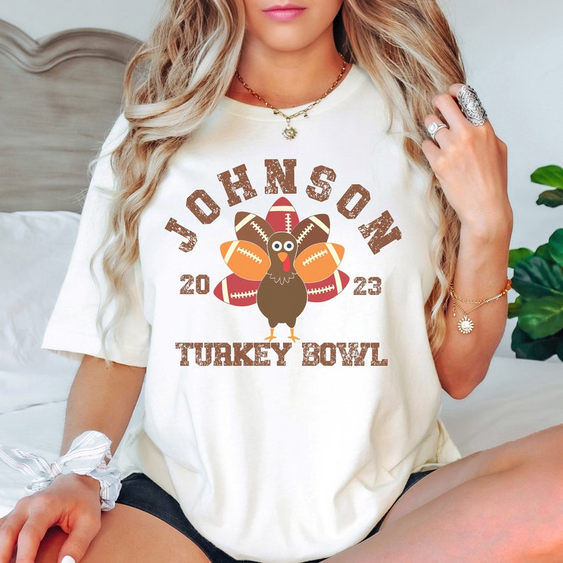 Op de afbeelding: Wit T-shirt met een bruine kalkoen-afbeelding die voetballen als veren draagt. De tekst "JOHNSON 20 23 TURKEY BOWL" is op het shirt gedrukt.