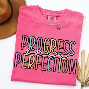 May include: A hot pink t-shirt with the words "PROGRESS OVER PERFECTION" in a colorful, outlined font. A gold necklace with a small pendant rests on the shirt. A brown hat and dried palm leaves are in the background.