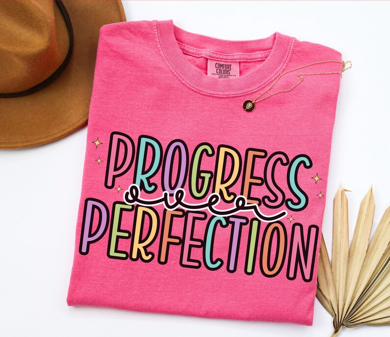 May include: A hot pink t-shirt with the words "PROGRESS OVER PERFECTION" in a colorful, outlined font. A gold necklace with a small pendant rests on the shirt. A brown hat and dried palm leaves are in the background.