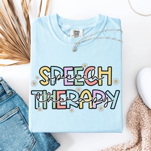 May include: Light blue t-shirt with the words "SPEECH THERAPY" in a colorful, playful font. The text is outlined in black and decorated with small daisy flowers. The shirt is folded on a white surface, next to a pair of blue jeans.