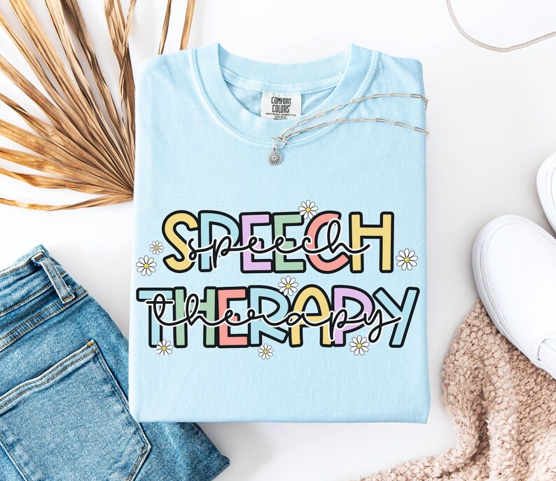 May include: Light blue t-shirt with the words "SPEECH THERAPY" in a colorful, playful font. The text is outlined in black and decorated with small daisy flowers. The shirt is folded on a white surface, next to a pair of blue jeans.