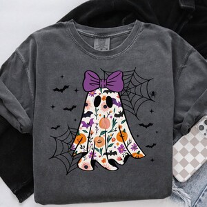 May include: Dark grey sweatshirt with a floral ghost design, a purple bow, pumpkins, and bats. The ghost has a floral pattern with pink, orange, and purple flowers. A spider web and spider are also included. A phone with a checkered case is visible.