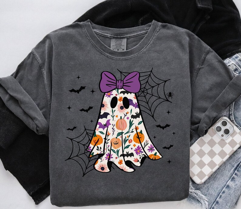 May include: Dark grey sweatshirt with a floral ghost design, a purple bow, pumpkins, and bats. The ghost has a floral pattern with pink, orange, and purple flowers. A spider web and spider are also included. A phone with a checkered case is visible.