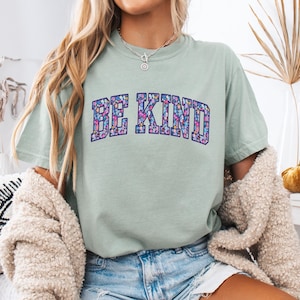 May include: A light green t-shirt with the words "BE KIND" in a floral design. The text is in a collegiate style font. The shirt is paired with denim shorts and a cream-colored cardigan.