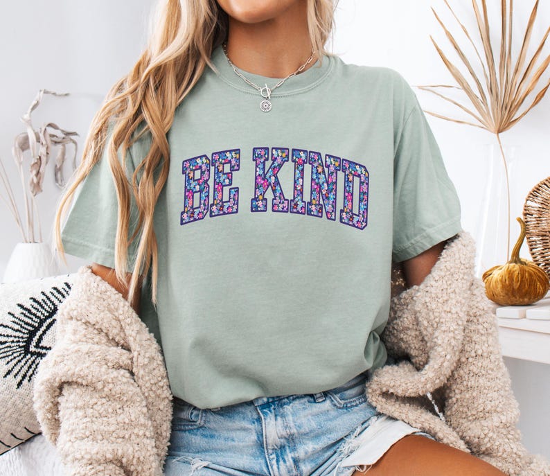 May include: A light green t-shirt with the words "BE KIND" in a floral design. The text is in a collegiate style font. The shirt is paired with denim shorts and a cream-colored cardigan.