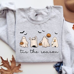 May include: A light gray sweatshirt featuring a Halloween design. The design includes four cartoon ghosts, bats, and the phrase "tis the season" in a script font. The ghosts are holding pumpkins and flowers.