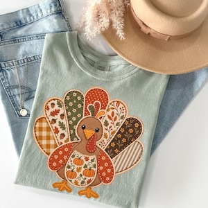 May include: A light sage green t-shirt featuring a central turkey graphic composed of various patterned fabric patches in orange, brown, and cream. The turkey design includes floral, plaid, acorn, and pumpkin prints, paired with light blue denim jeans.