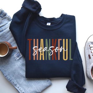 Puede incluir: Sudadera azul marino con el texto "Thankful Season" en rayas verticales de colores. El texto est&aacute; en blanco.