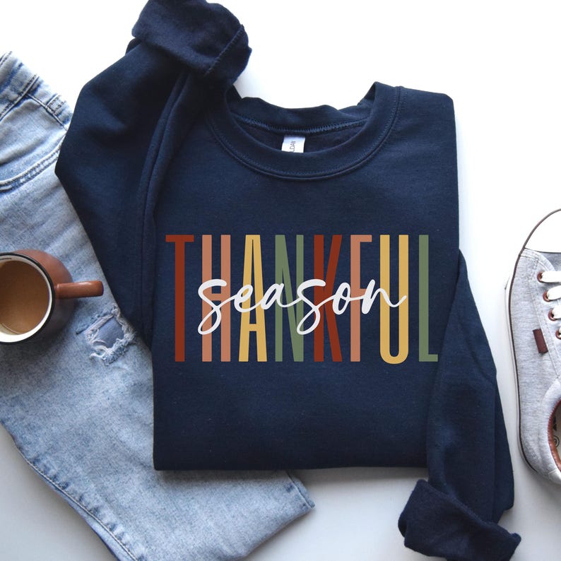 Puede incluir: Sudadera azul marino con el texto "Thankful Season" en rayas verticales de colores. El texto est&aacute; en blanco.